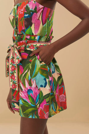 A person wears the Farm Rio Jardin De Reve Multicolor Romper, featuring a floral print, self-tie waist, and pockets, photographed against a beige background.