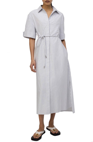 A person wearing a Simkhai Grady Dress featuring a long white and gray striped design with a belted waist, rolled-up sleeves, and black flip-flops with white straps—a perfect blend of modern sophistication.