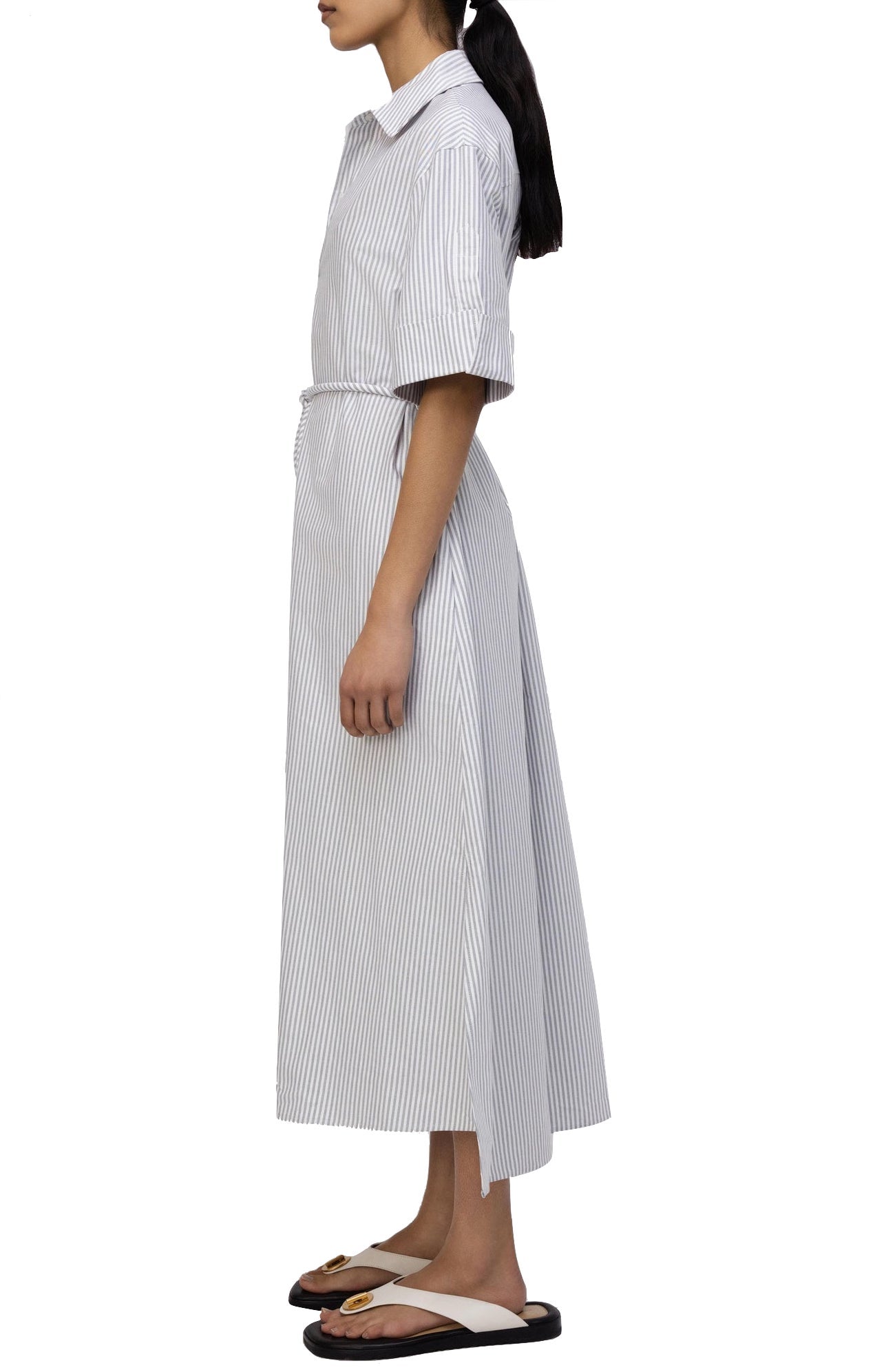 Side view of a person wearing the Simkhai Grady Dress, an ensemble of modern sophistication featuring a white and light grey striped pattern with short sleeves, a belted waist, and black and white slide sandals.