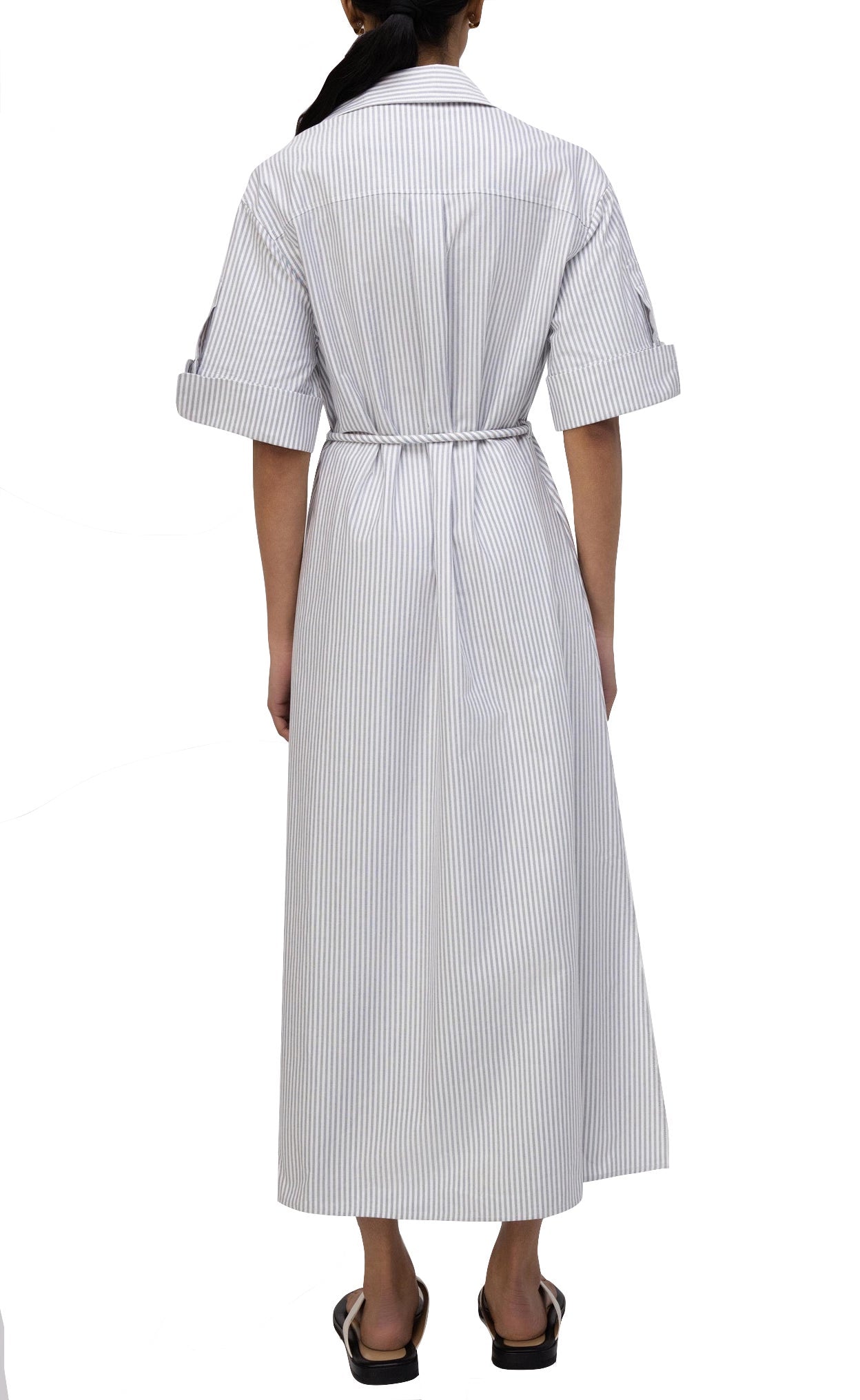 A person stands facing away, wearing the Simkhai Grady Dress—a long, light-colored striped design with rolled-up sleeves and a belted waist—paired with black sandals, exuding modern sophistication.