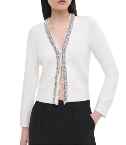 A woman models the Simkhai Lieghton Boucle Cardigan, highlighting its flattering fit with silver trim as it layers perfectly over her black pants, while she stands with one hand in her pocket.