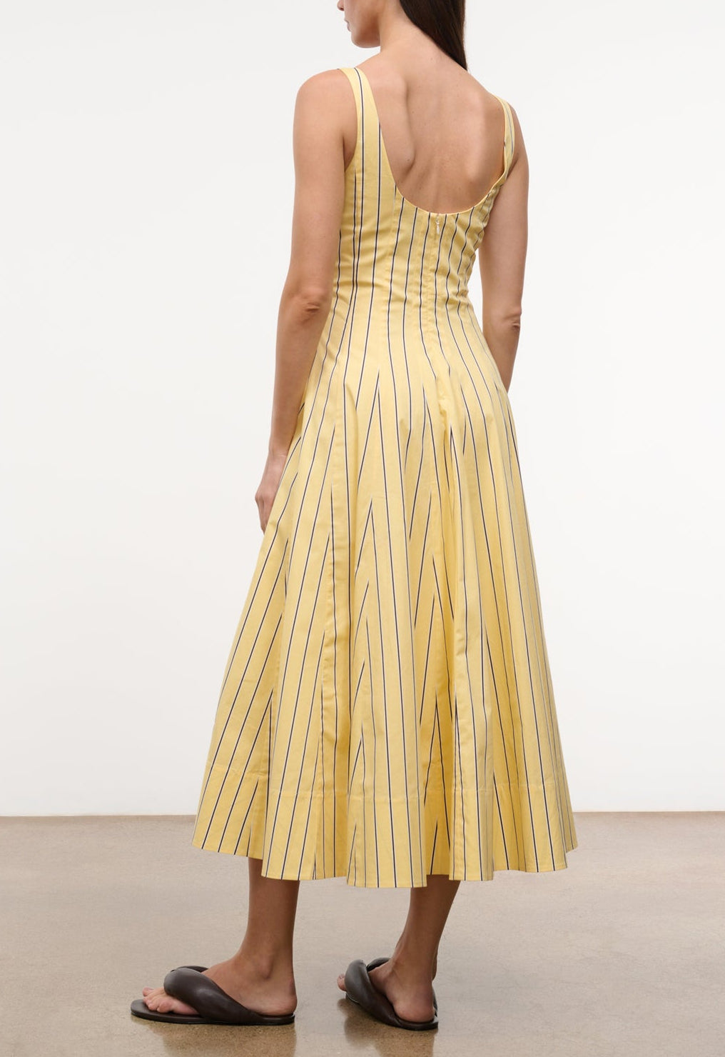 A woman stands on a light brown floor wearing the Staud Wells Monday Stripe Lobster Dress—a yellow, sleeveless, mid-length fitted waist dress with black vertical stripes—and black slide sandals.