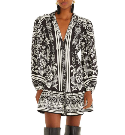 A person wears the Farm Rio Lisbon Black Mini Dress featuring bold botanical prints, long sleeves, a v-neckline, and pairs it with knee-high black boots for a chic look.