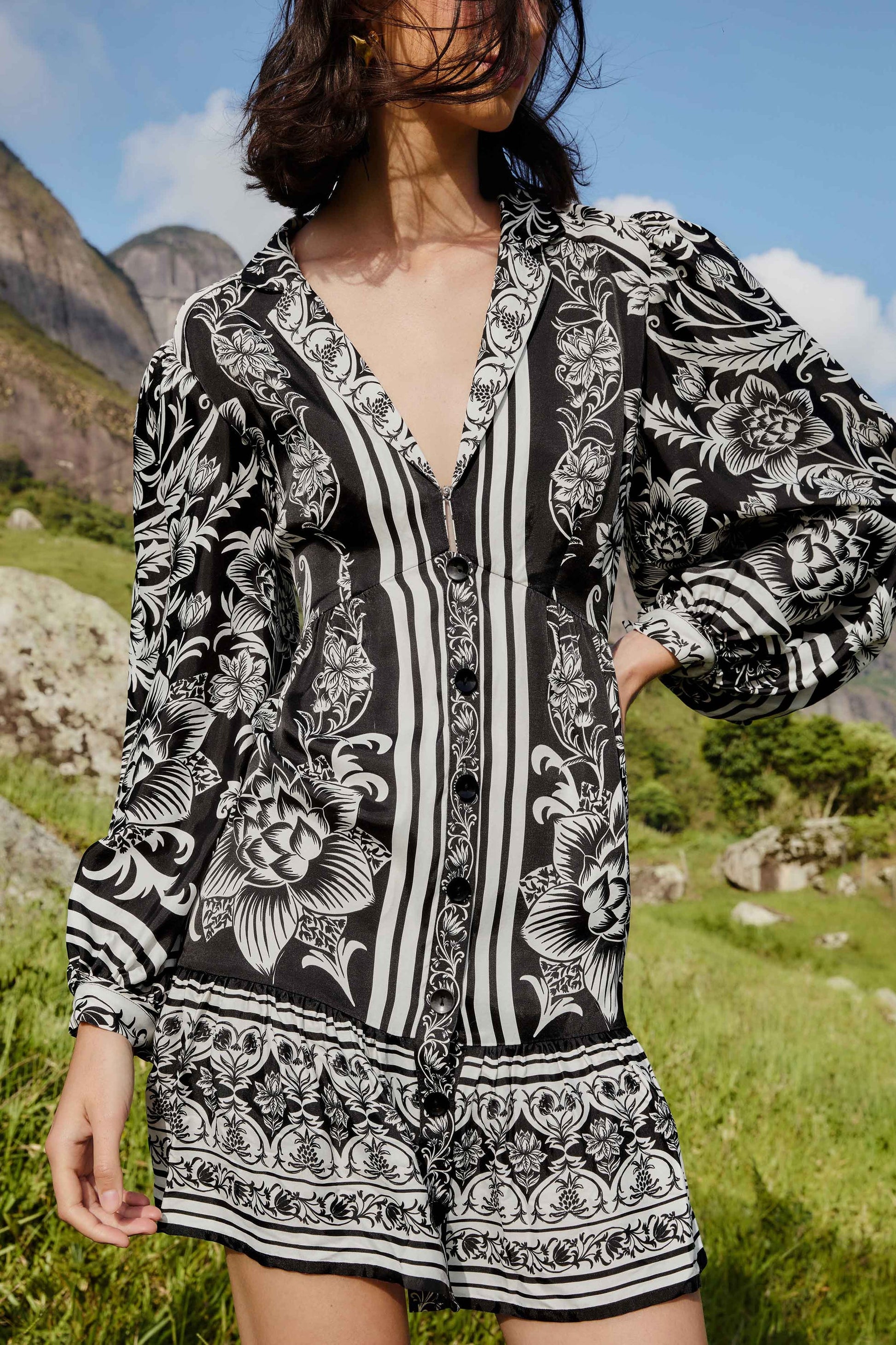 A person stands outdoors with hills and blue sky behind them, wearing the Farm Rio Lisbon Black Mini Dress—a long-sleeve, v-neck mini dress featuring bold botanical prints.