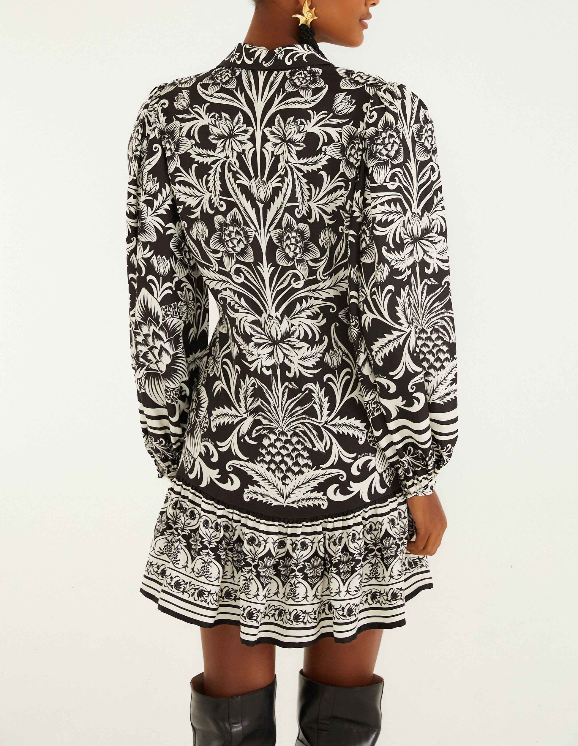 Someone wearing the Farm Rio Lisbon Black Mini Dress, a lightweight floral piece with long sleeves and a ruffled hem, stands with their back to the camera.