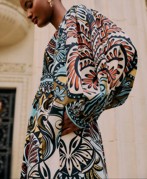 Wearing the vibrant Balamana Maxi Dress by Farm Rio, a person stands before an ornate, pale building, capturing laid-back tropical getaway vibes with its bold patterns and wide sleeves.