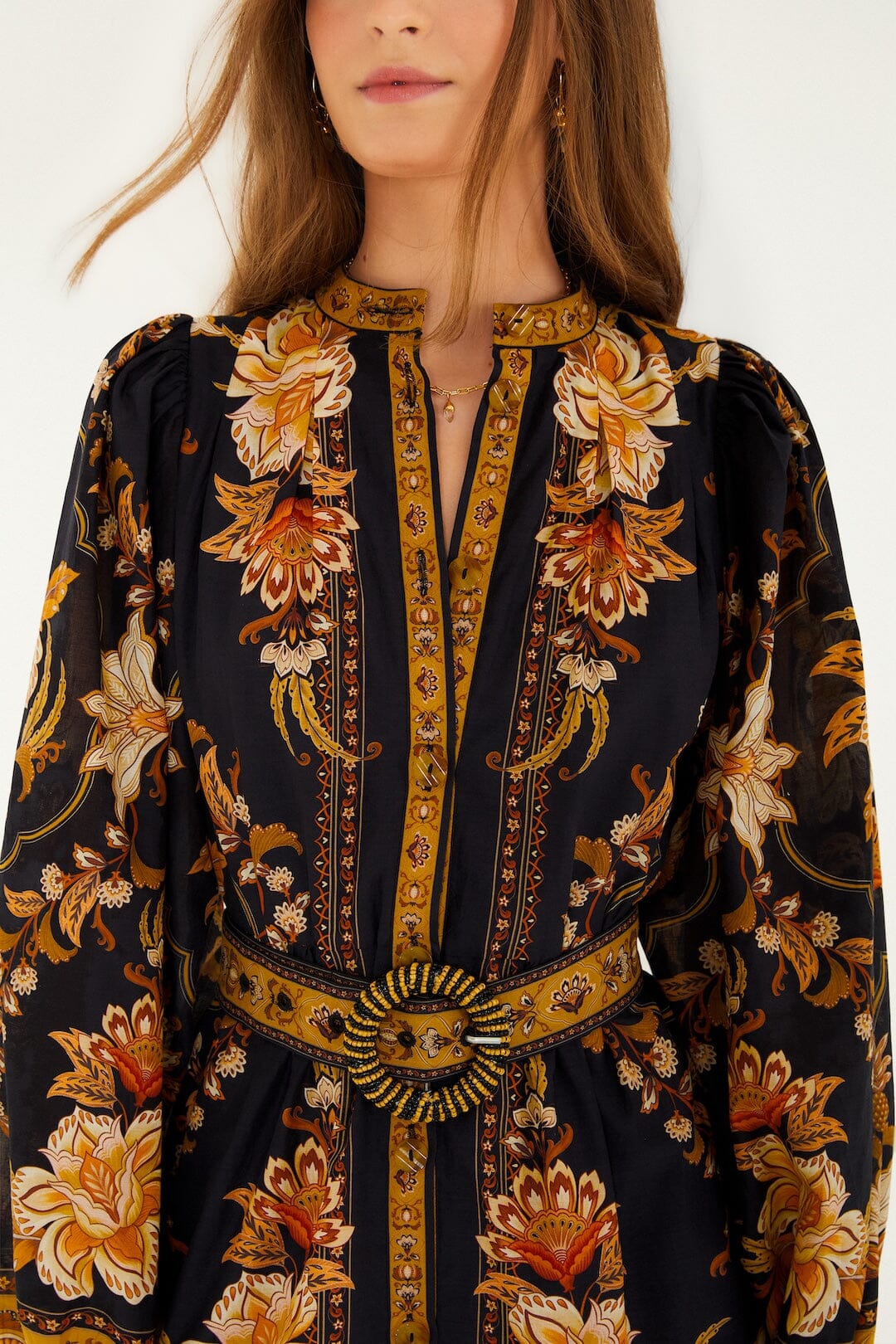 A woman wears the Majestic Garden Black Mini Dress by Farm Rio, featuring gold, orange, and cream floral patterns, a beaded buckle belt, and only part of her face visible.
