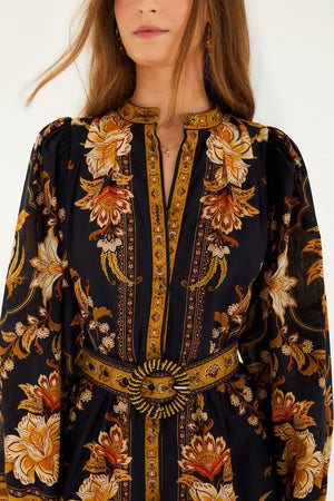 A woman wears the Majestic Garden Black Mini Dress by Farm Rio, featuring gold, orange, and cream floral patterns, a beaded buckle belt, and only part of her face visible.