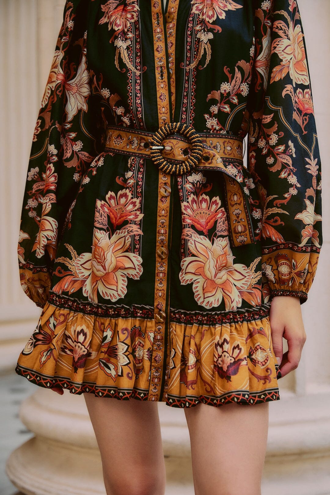 Person wearing the Farm Rio Majestic Garden Black Mini Dress, a long-sleeved floral mini with a beaded buckle belt and ruffled hem, shown from shoulders to mid-thighs.