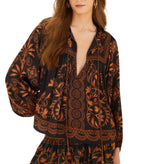 A woman poses against a white background wearing the Farm Rio Cornely Ainika Blouse, a black and gold floral top with beaded tassel ties, paired with a matching skirt.