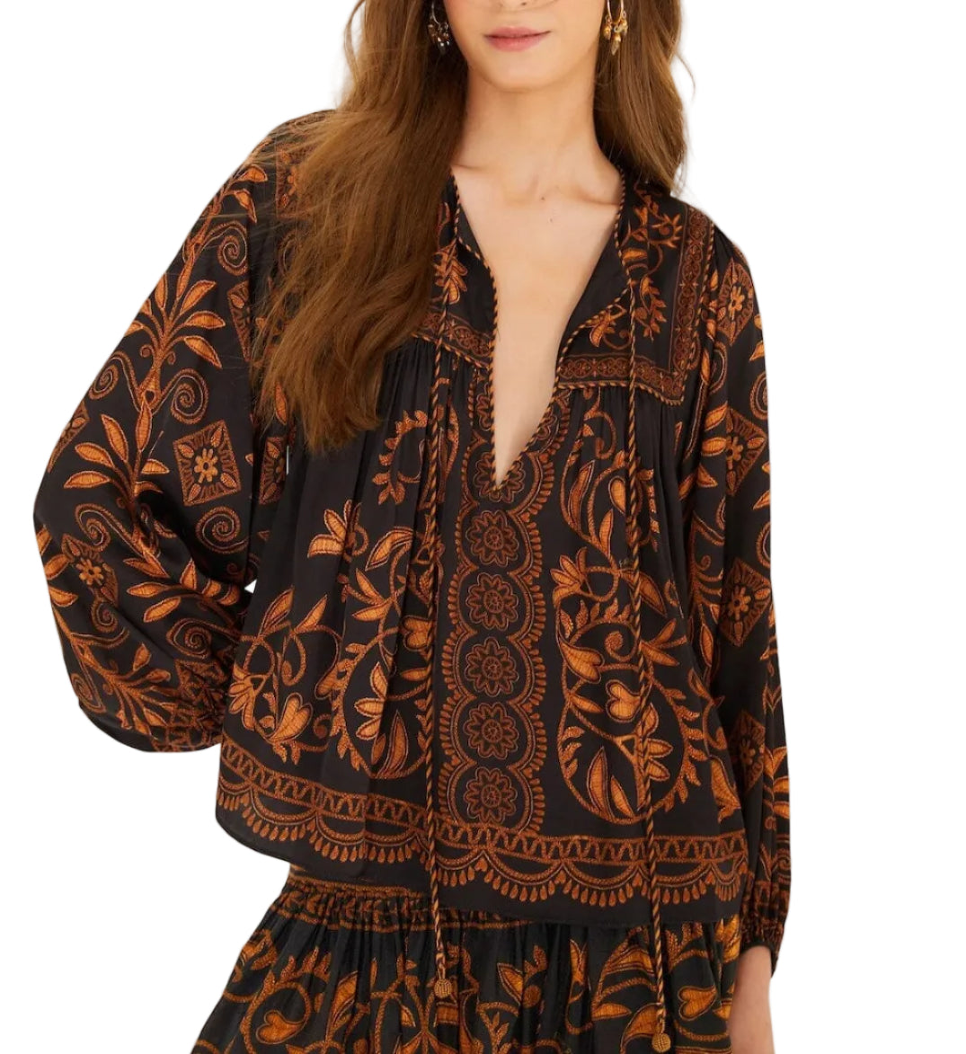 A woman poses against a white background wearing the Farm Rio Cornely Ainika Blouse, a black and gold floral top with beaded tassel ties, paired with a matching skirt.