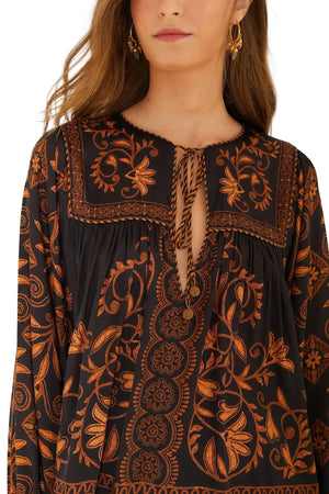 A woman wears the Cornely Ainika Blouse by Farm Rio—a black and gold floral top with intricate embroidery and beaded tassel ties at the neckline.