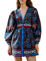 A woman wears the Farm Rio Latina Scarf Mini Dress, featuring blue, red, and white floral patterns, long puff sleeves, a deep V-neckline, and chic front buttons.