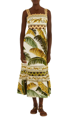 A person wearing Farm Rio’s Trancoso Sand Sleeveless Maxi Dress—an eco-friendly, ankle-length style in a green and yellow leaf print with geometric trim—paired with brown sandals.