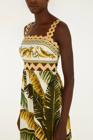 A woman wears the Farm Rio Trancoso Sand Sleeveless Maxi Dress, an eco-friendly piece featuring a scalloped neckline and a banana leaf and fruit print in green, yellow, and beige tones.