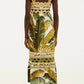 A woman faces away, wearing the Farm Rio Trancoso Sand Sleeveless Maxi Dress—an ankle-length, eco-friendly Lenzing fiber dress with green and yellow tropical leaf print and scalloped trim—paired with brown flat sandals.