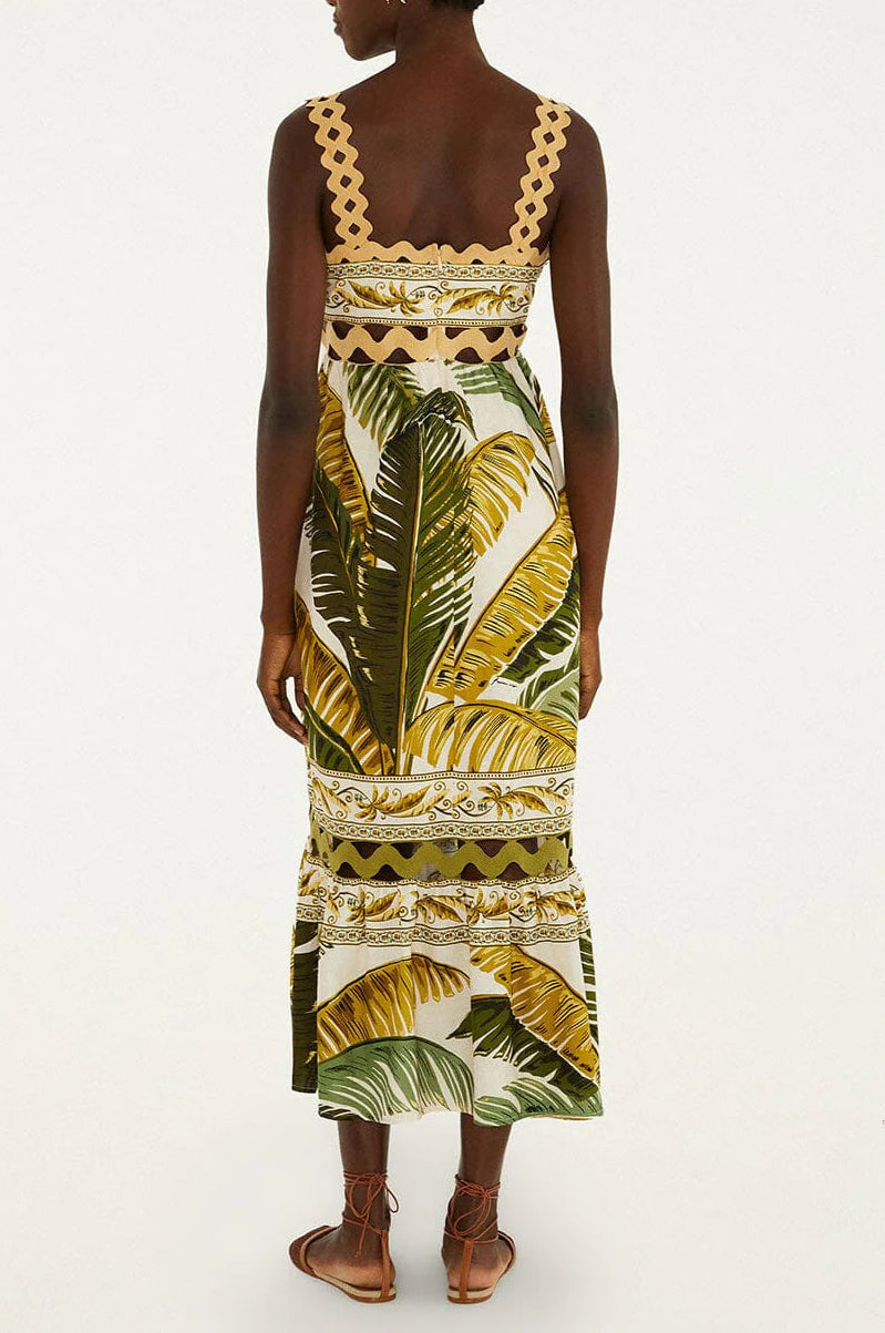 A woman faces away, wearing the Farm Rio Trancoso Sand Sleeveless Maxi Dress—an ankle-length, eco-friendly Lenzing fiber dress with green and yellow tropical leaf print and scalloped trim—paired with brown flat sandals.