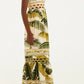 A person stands sideways wearing the Farm Rio Trancoso Sand Sleeveless Maxi Dress, featuring a green and yellow leaf pattern. The eco-friendly, ankle-length dress is paired with brown sandals against a plain background.