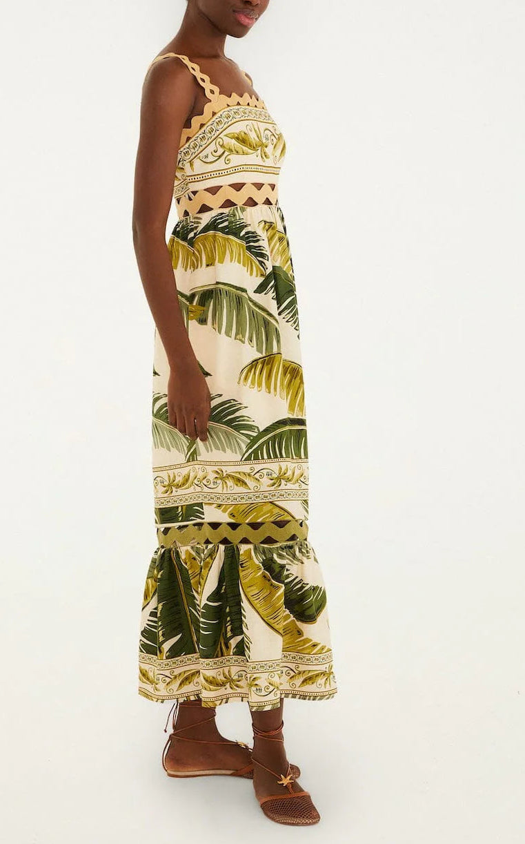 A person stands sideways wearing the Farm Rio Trancoso Sand Sleeveless Maxi Dress, featuring a green and yellow leaf pattern. The eco-friendly, ankle-length dress is paired with brown sandals against a plain background.