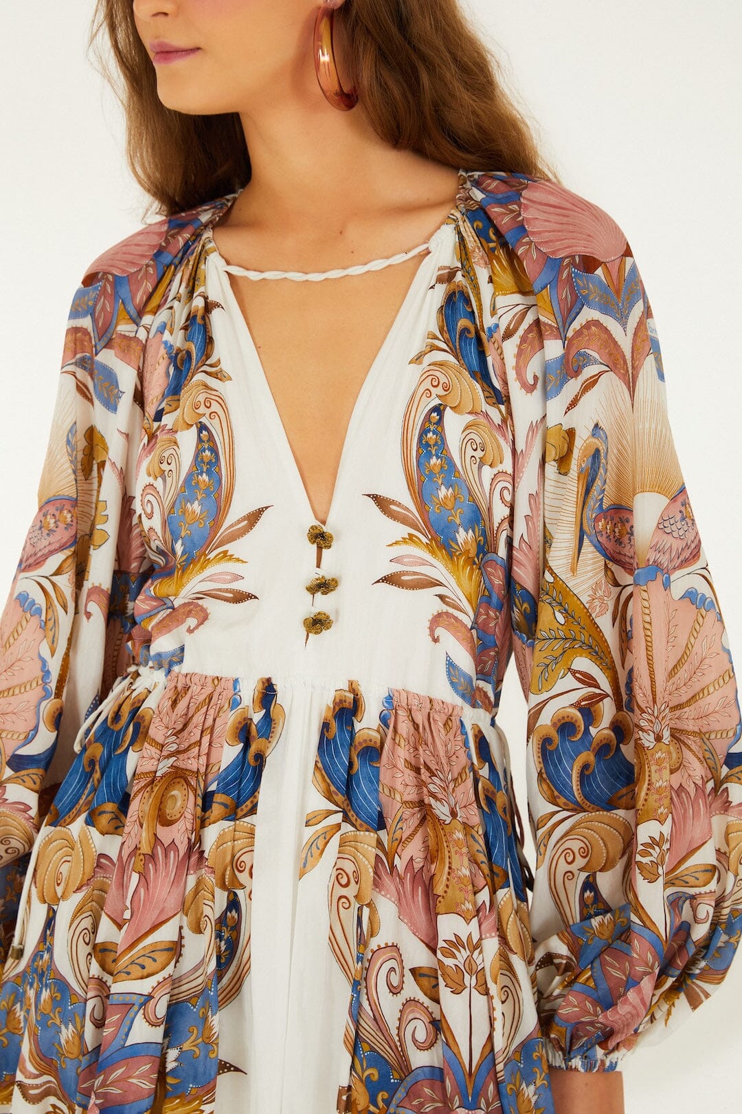 A woman wears the Farm Rio Sea Garden Off White Mini Bubble Dress, featuring long sleeves, a deep V-neck, rope detail, and a colorful blue, pink, and gold floral-paisley pattern.