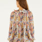 A woman with long brown hair is seen from the back wearing a Farm Rio Sea Garden Off White Long Sleeve Blouse with floral patterns and ruffles, paired with brown shorts.