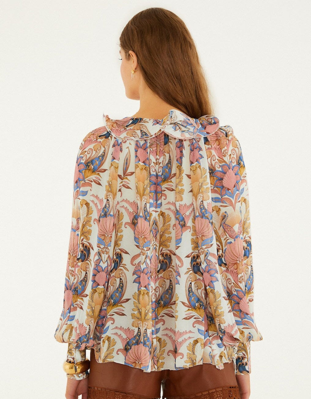 A woman with long brown hair is seen from the back wearing a Farm Rio Sea Garden Off White Long Sleeve Blouse with floral patterns and ruffles, paired with brown shorts.