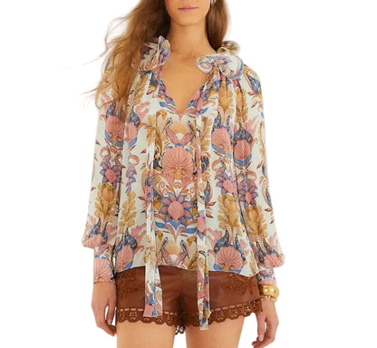 A woman wears the Farm Rio Sea Garden Off White Long Sleeve Blouse with ruffles and a bow at the collar, paired with brown lace-trimmed shorts.