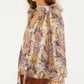A woman wears the Farm Rio Sea Garden Off White Long Sleeve Blouse with ruffled sleeves and a neck bow, paired with a brown embroidered skirt and a gold bangle.