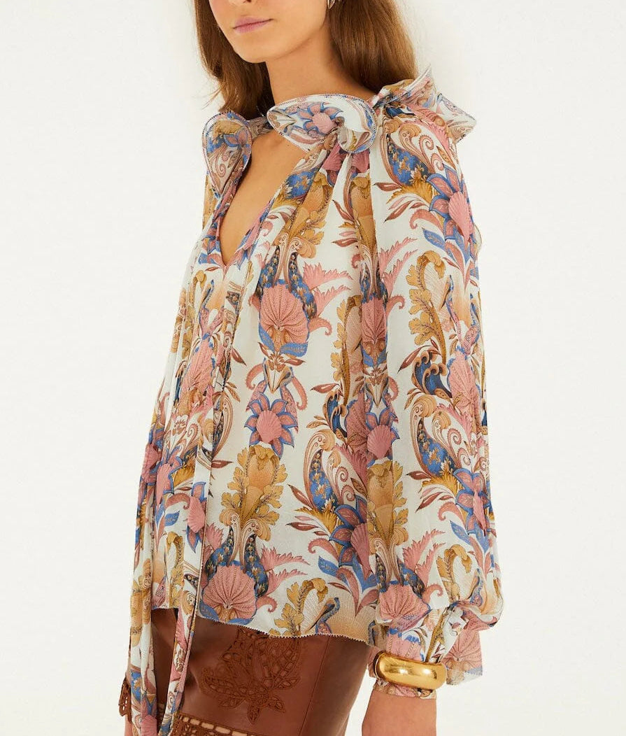 A woman wears the Farm Rio Sea Garden Off White Long Sleeve Blouse with ruffled sleeves and a neck bow, paired with a brown embroidered skirt and a gold bangle.