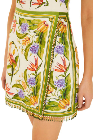 A close-up of the Farm Rio Sand Tropical Scarf Sleeveless Mini Dress, showcasing its vibrant floral print, green borders, and bead fringe detailing along the hem.