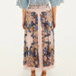 A woman faces away, wearing a blue scalloped top and the Farm Rio Sea Garden Scarf Soft Pink Maxi Skirt with intricate blue and beige floral patterns, paired with brown sandals.