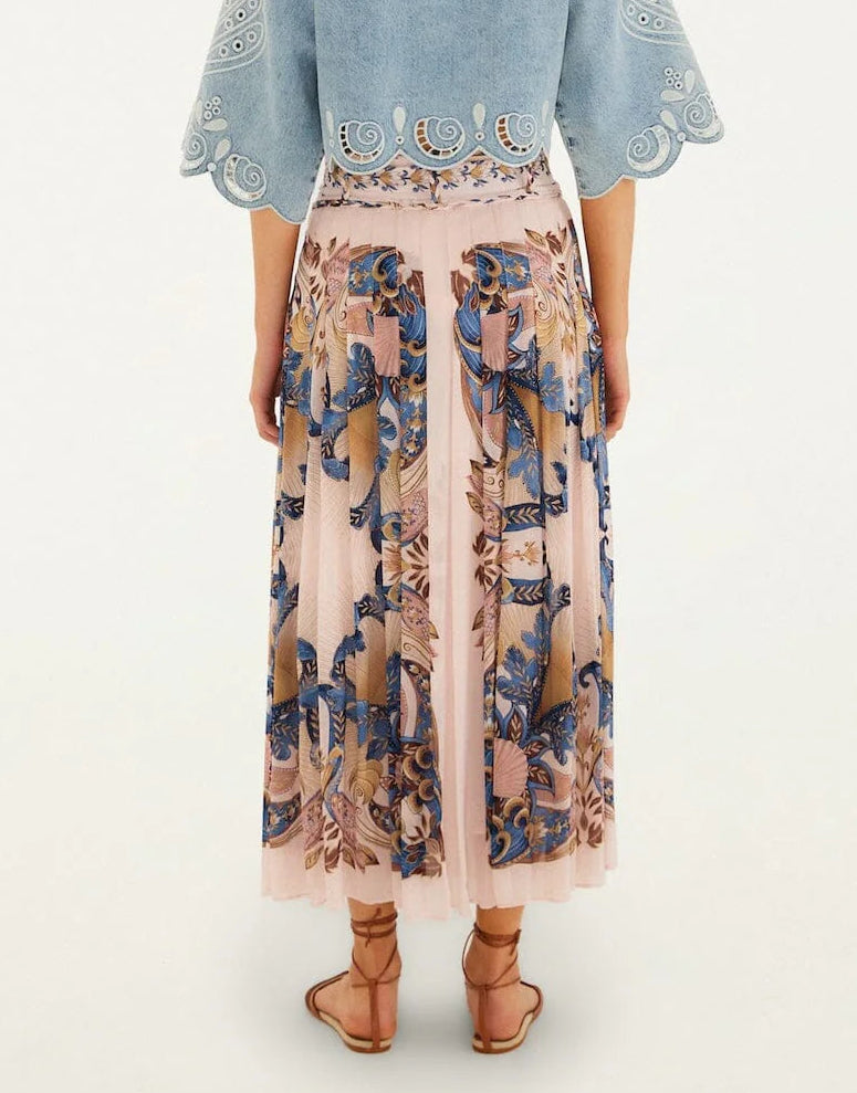 A woman faces away, wearing a blue scalloped top and the Farm Rio Sea Garden Scarf Soft Pink Maxi Skirt with intricate blue and beige floral patterns, paired with brown sandals.