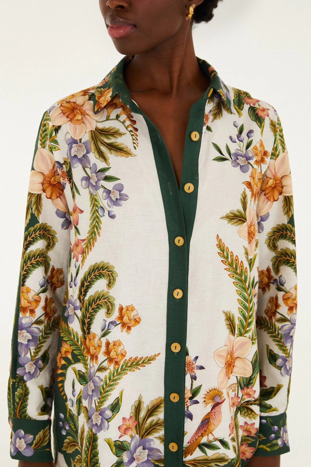 A person wears the Farm Rio Nature Sand Shirt—a premium linen collared button-down with green trim and vibrant floral and bird prints on a white background.