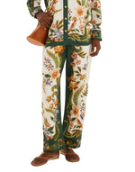 A person wears Farm Rio’s Nature Sand Pants and a matching floral shirt with green accents, paired with a brown bag and brown flat shoes.