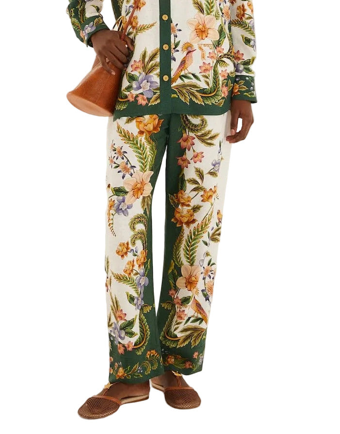A person wears Farm Rio’s Nature Sand Pants and a matching floral shirt with green accents, paired with a brown bag and brown flat shoes.