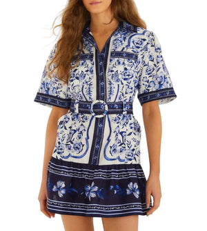 A woman wears the Porcelain Garden Off White Mini Dress by Farm Rio, a short-sleeve, collared linen dress with blue and white floral patterns, dark blue trim, and a belted waist for a breezy, breathable look.