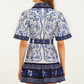 A woman stands facing away, wearing the Porcelain Garden Off White Mini Dress by Farm Rio—a short-sleeved, floral-patterned linen dress with a matching belt, combining elegance with breathable comfort.