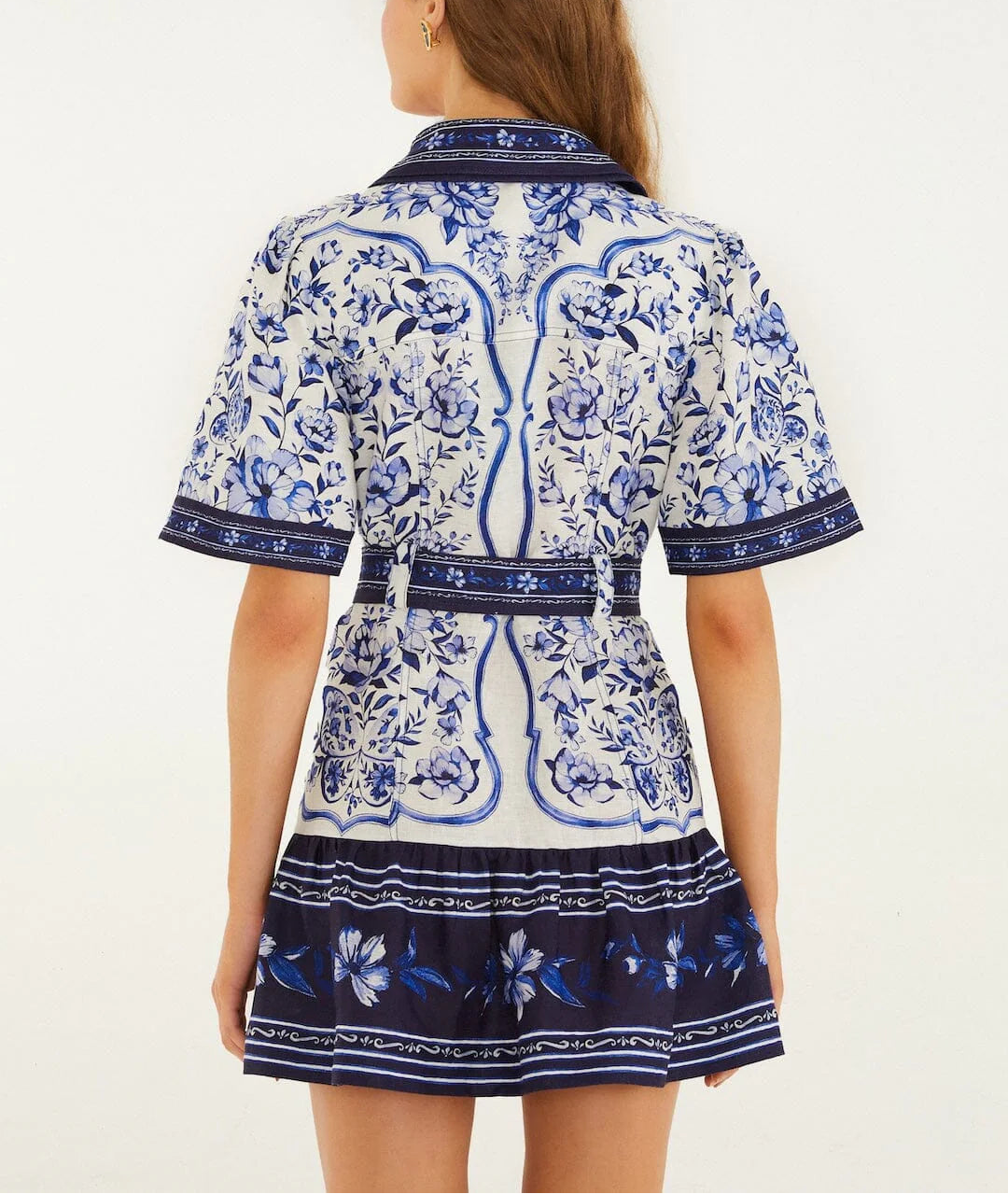 A woman stands facing away, wearing the Porcelain Garden Off White Mini Dress by Farm Rio—a short-sleeved, floral-patterned linen dress with a matching belt, combining elegance with breathable comfort.
