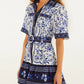 A woman wears the Porcelain Garden Off White Mini Dress by Farm Rio, a short-sleeved, belted linen dress with blue floral patterns and dark blue bands at the hem and cuffs.
