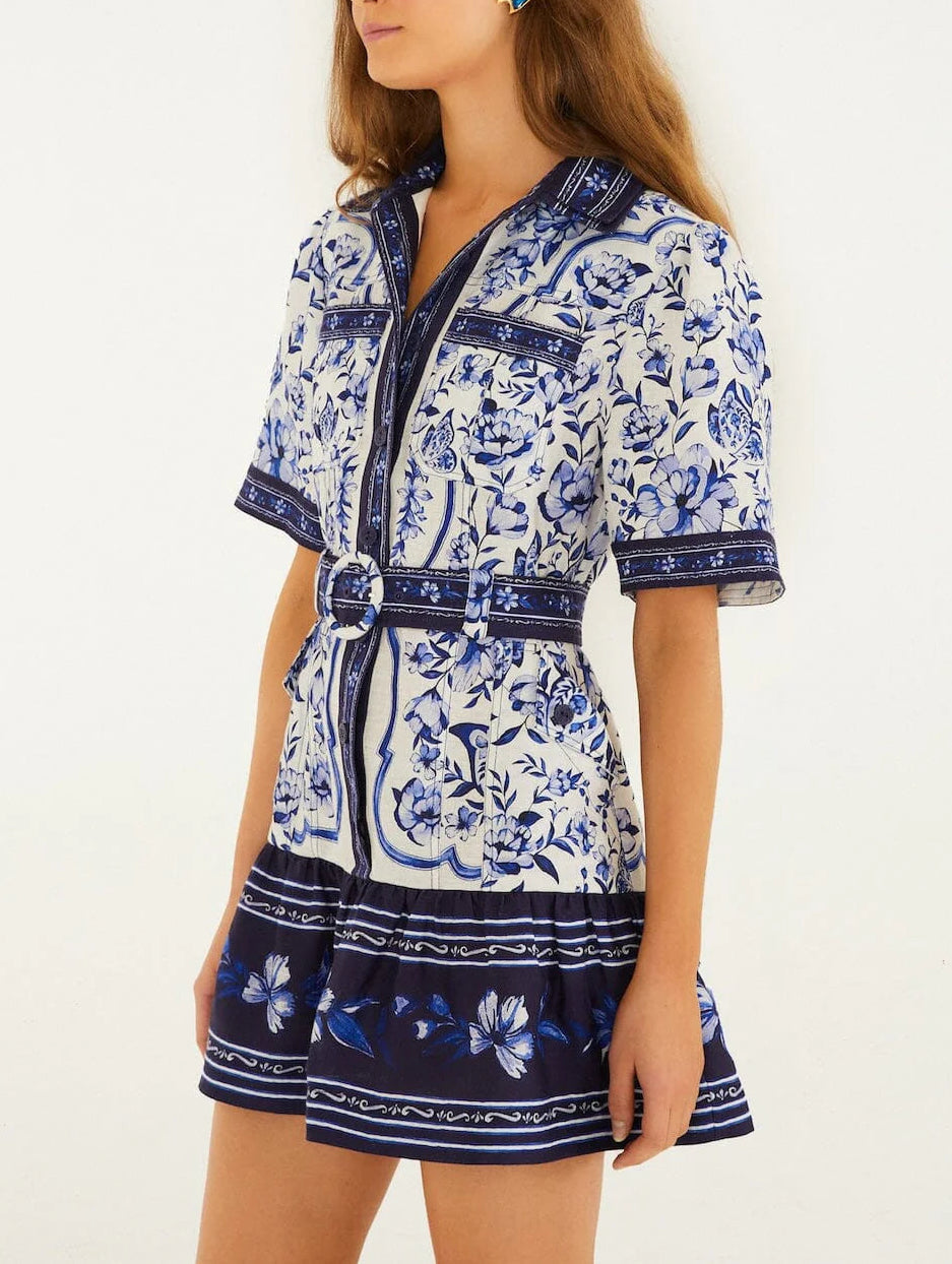 A woman wears the Porcelain Garden Off White Mini Dress by Farm Rio, a short-sleeved, belted linen dress with blue floral patterns and dark blue bands at the hem and cuffs.