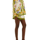 A person in Farm Rio’s Sand Tropical Scarf Shorts and a matching floral-print jacket with green trim, paired with green heeled mules, stands against a white background—a tropical-inspired summer look.