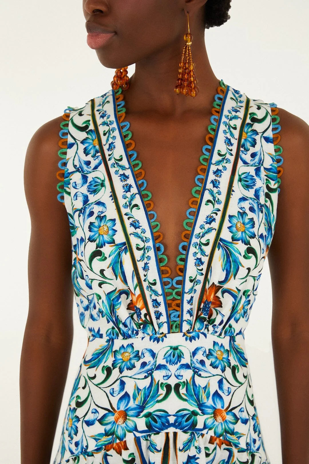 A person wearing the Farm Rio Flora Tile Off White Midi Dress, featuring a sleeveless V-neck, blue and green floral tile print, scalloped hem, and orange drop earrings.