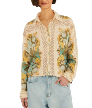 A woman wears the Farm Rio Royal Sea Crochet Detail Shirt, a light-colored button-up with a floral and bird print in an Ecovero blend, paired with light blue jeans and standing with one hand in her pocket.