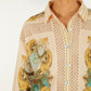 A woman wears the Royal Sea Crochet Detail Shirt by Farm Rio—a patterned button-up with gold, blue, and beige designs in an Ecovero blend. Featuring a collar and visible buttons, it's ideal for lightweight layering. Her face is partially out of frame.