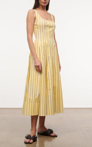 A woman wears the Staud Wells Monday Stripe Lobster Dress—a sleeveless, yellow cotton poplin dress with vertical black stripes—and black sandals, standing on a smooth light brown floor against a plain white background.