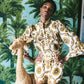 A person stands indoors wearing the Cara Cara Elowen Top with ruffled long sleeves, hands on hips, beside a wicker giraffe and palm tree wallpaper.