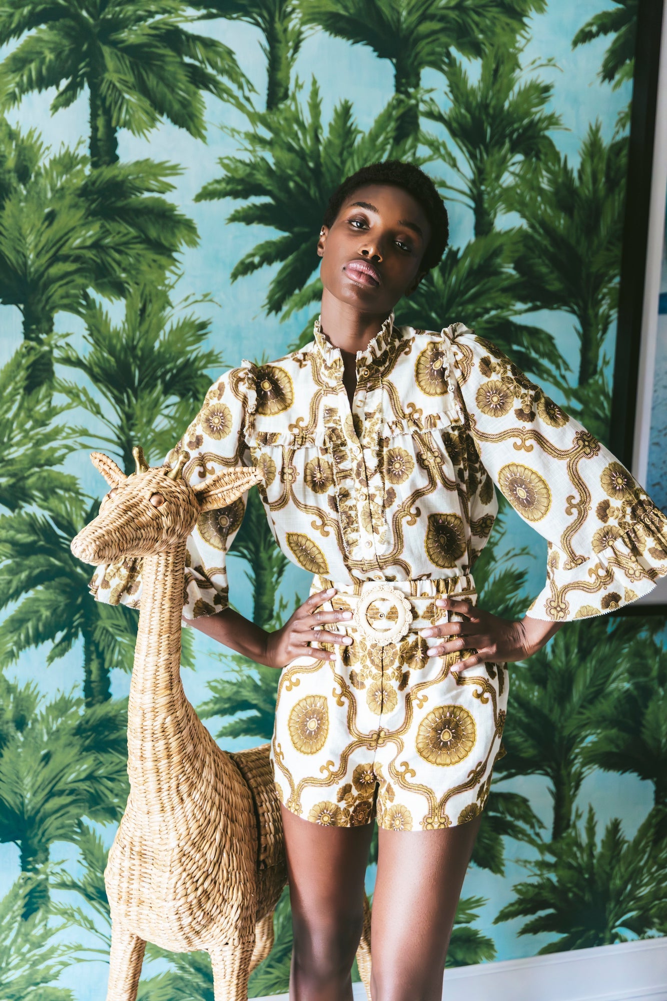 A person stands indoors wearing the Cara Cara Elowen Top with ruffled long sleeves, hands on hips, beside a wicker giraffe and palm tree wallpaper.
