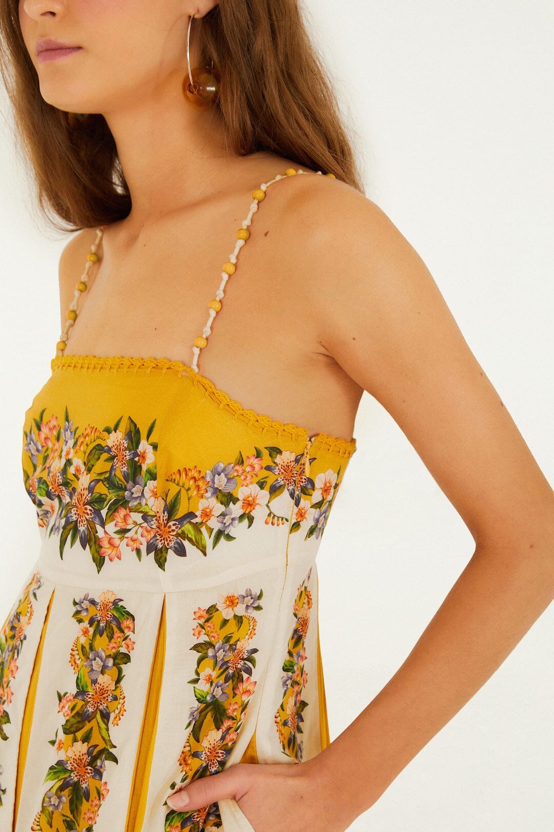 A woman wearing the Farm Rio Sand Hilo Stripes Dress with beaded straps and floral patterns stands against a plain light background, accessorized with large hoop earrings.