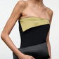 A woman in a strapless black dress with an olive green accent holds the Staud Muse Clutch, an elegant evening accessory, against a plain background.