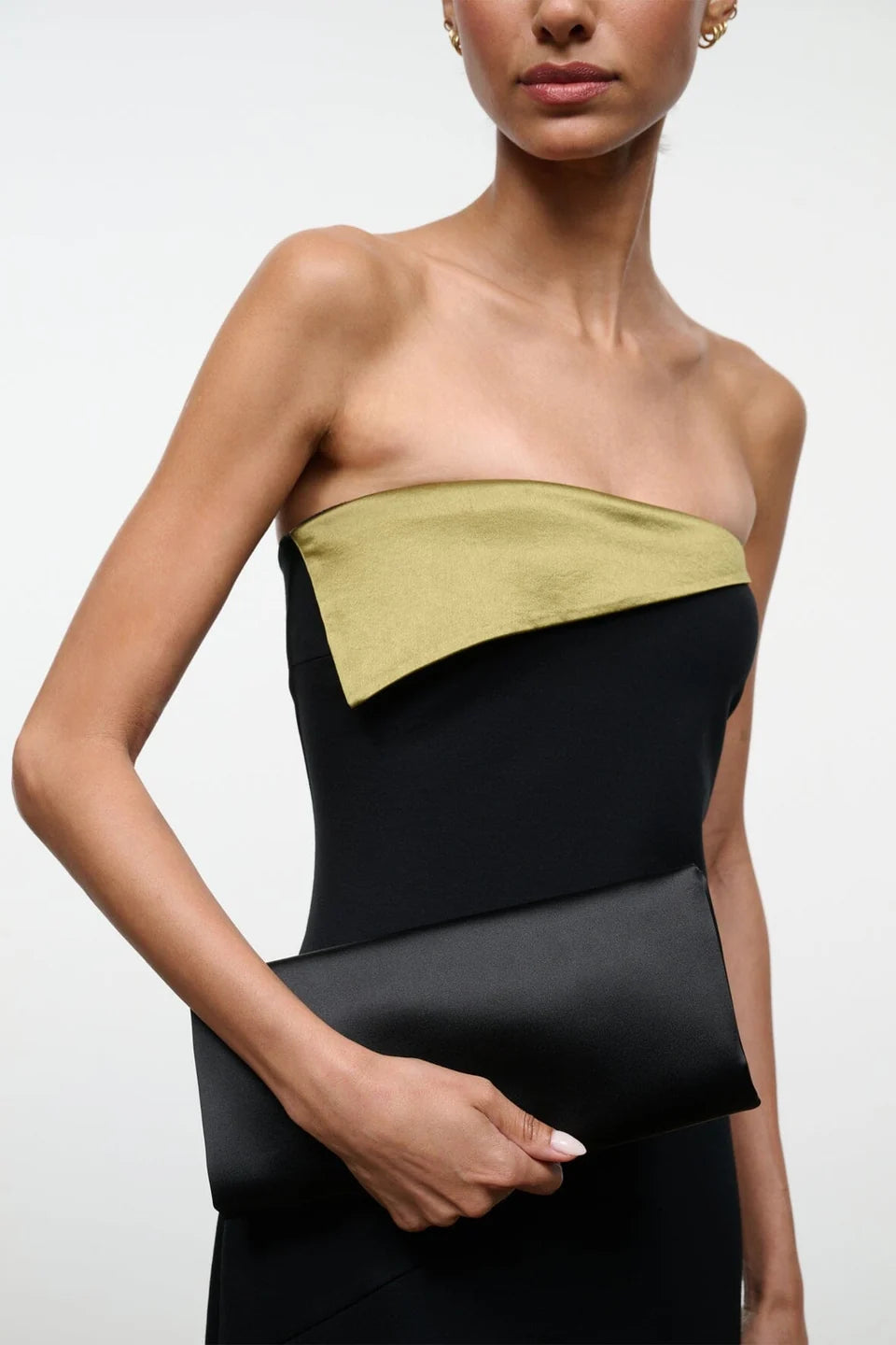 A woman in a strapless black dress with an olive green accent holds the Staud Muse Clutch, an elegant evening accessory, against a plain background.